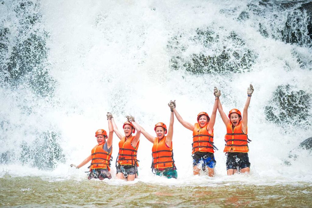 Eine Gruppe ausländischer Reisender, alle mit Schwimmwesten, steht mit erhobenen Händen vor einem Wasserfall während einer Canyoning-Tour in Dalat