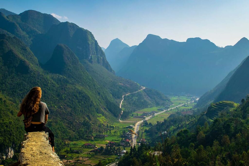 Frau sitzt auf der Kante eines Felsens und genießt die atemberaubende Aussicht auf die Berge entlang des Ha Giang Loop