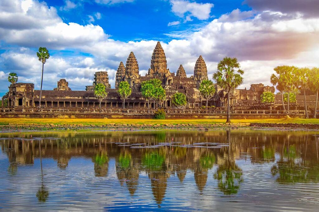 Der Tempelkomplex von Angkor Wat in Kambodscha spiegelt sich im Wasser davor, umgeben von Palmen unter einem bewölkten Himmel