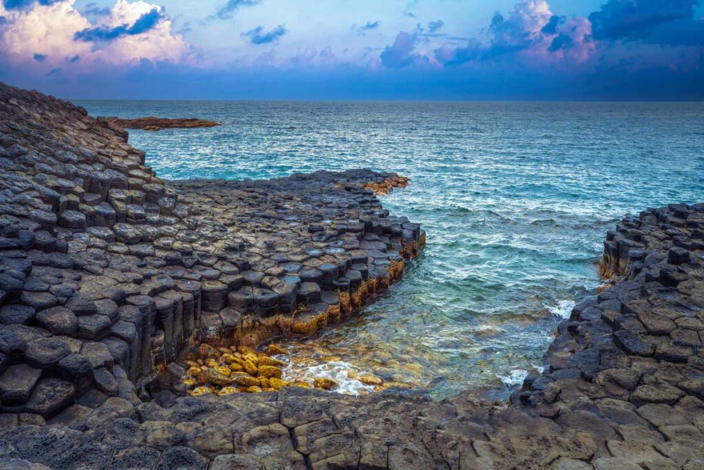 Sechseckige Basaltsäulen des Da Dia Riffs in Phu Yen, entstanden durch abgekühltes Lavagestein, mit Wellen, die bei bewölktem Sonnenuntergang gegen die Felsformationen schlagen — ein geologisch besonderes Phänomen an der Küste von Tuy Hoa