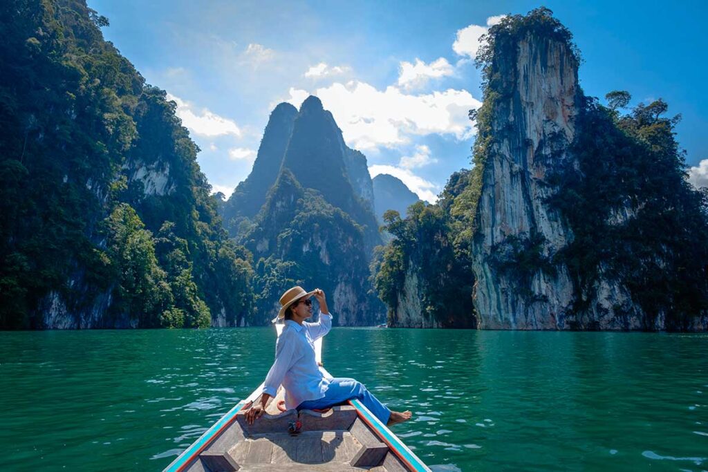 Frau sitzt auf dem Bug eines Longtailboots auf dem grünen Wasser des Cheow Lan Lake im Khao-Sok-Nationalpark, umgeben von hohen Kalksteinfelsen mit tropischem Bewuchs