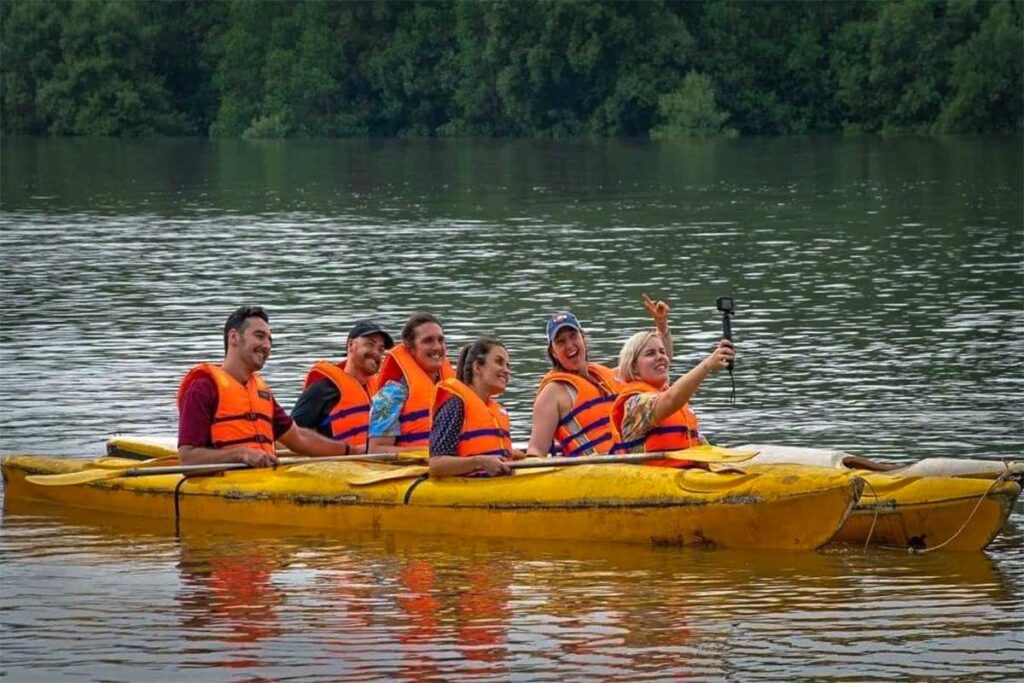 Gruppe von Reisenden in orangefarbenen Schwimmwesten beim Kajakfahren in einem großen gelben Kajak auf dem Fluss bei Dan Xay im Can Gio Reservat, mit Mangrovenwald im Hintergrund — Kajakfahren ist eine der aktiveren Möglichkeiten, das Reservat zu erkunden