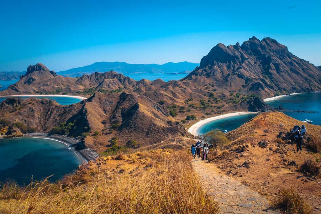 Gruppe von Wanderern auf einem Steinpfad über die kargen Hügel von Padar Island bei Flores in Indonesien, mit drei Buchten und weißen Stränden im Hintergrund sichtbar
