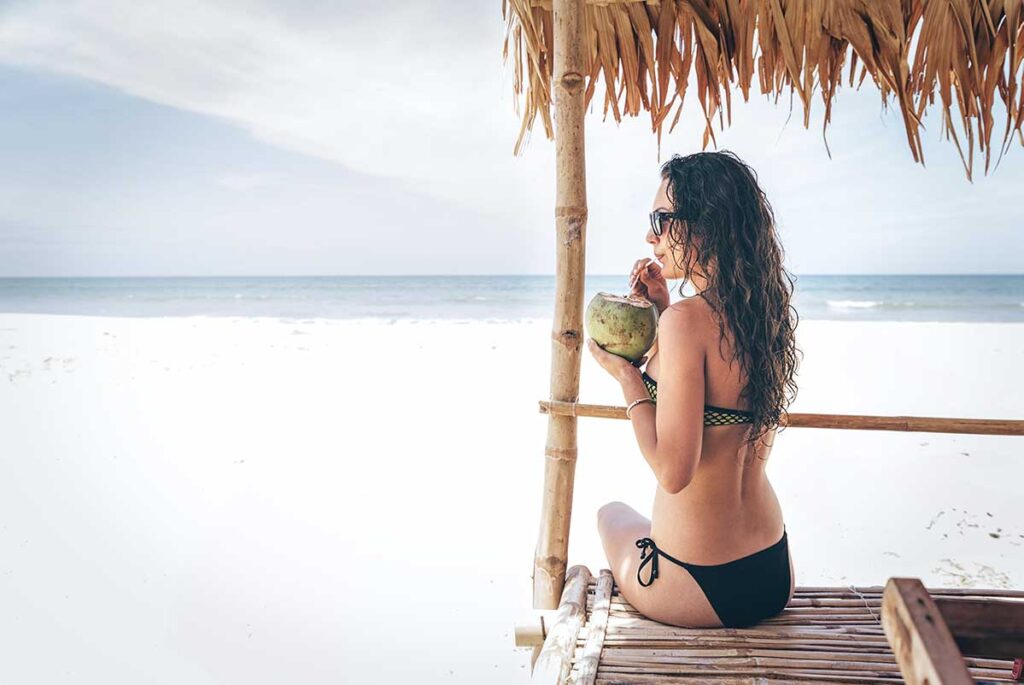 Frau im schwarzen Bikini sitzt unter einer Bambus-Strandhütte an einem weißen Sandstrand in Vietnam und trinkt aus einer frischen Kokosnuss mit Sonnenbrille
