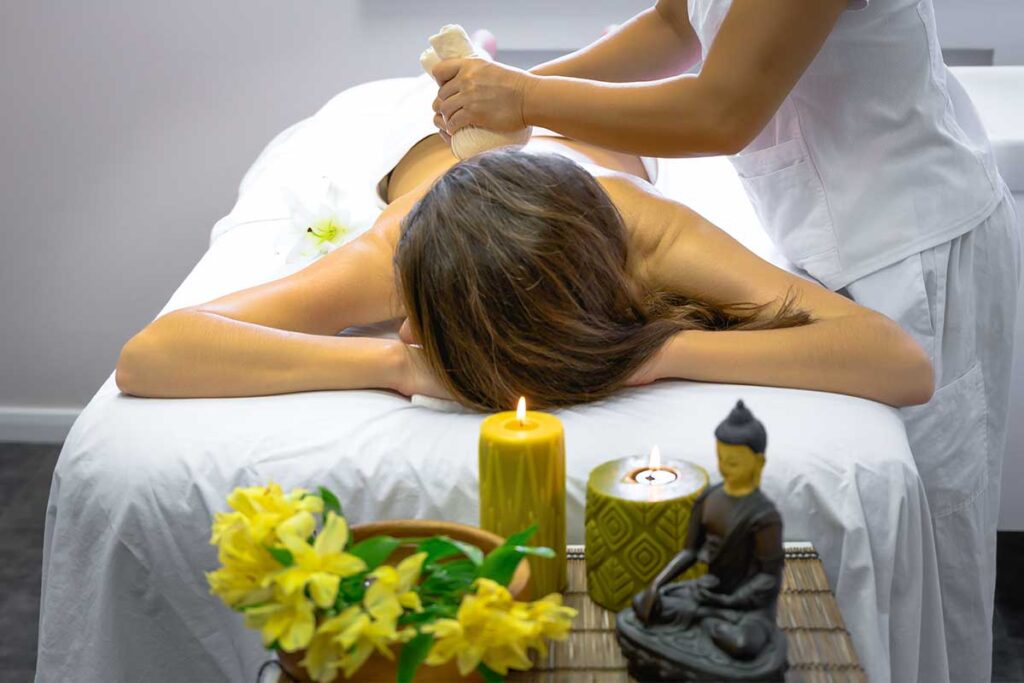 Frau erhält eine Ganzkörpermassage mit Kräuterstempel auf dem Rücken in einem vietnamesischen Spa, mit Kerzen und einer Buddhastatue im Vordergrund