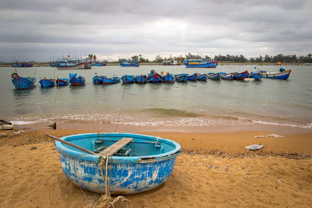Kleiner Fischerhafen in Dong Tac bei Tuy Hoa mit einem runden Korbboot im Sand und Dutzenden blauer Fischerboote vor Anker im trüben Wasser unter bewölktem Himmel