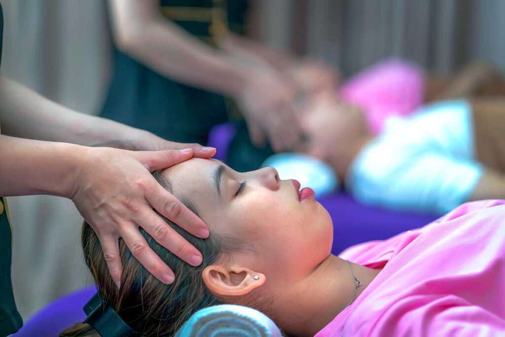 Vietnamesischer Therapeut gibt einer Frau in einem Massagesalon in Vietnam eine Kopfmassage, mit einem zweiten Kunden im Hintergrund sichtbar