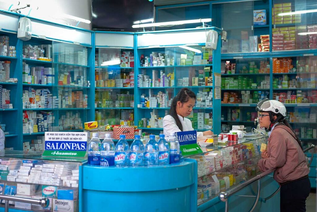 Lokale Apotheke in Vietnam mit einer Mitarbeiterin im weißen Kittel hinter dem Tresen, die einen Kunden mit Motorradhelm bedient, umgeben von Regalen voller Medikamente