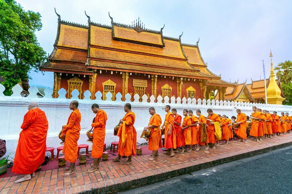 luang prabang laos tempel kultur 1 Reihe buddhistischer Mönche in orangefarbenen Gewändern beim täglichen Almosengang entlang eines goldenen Tempels in Luang Prabang, Laos