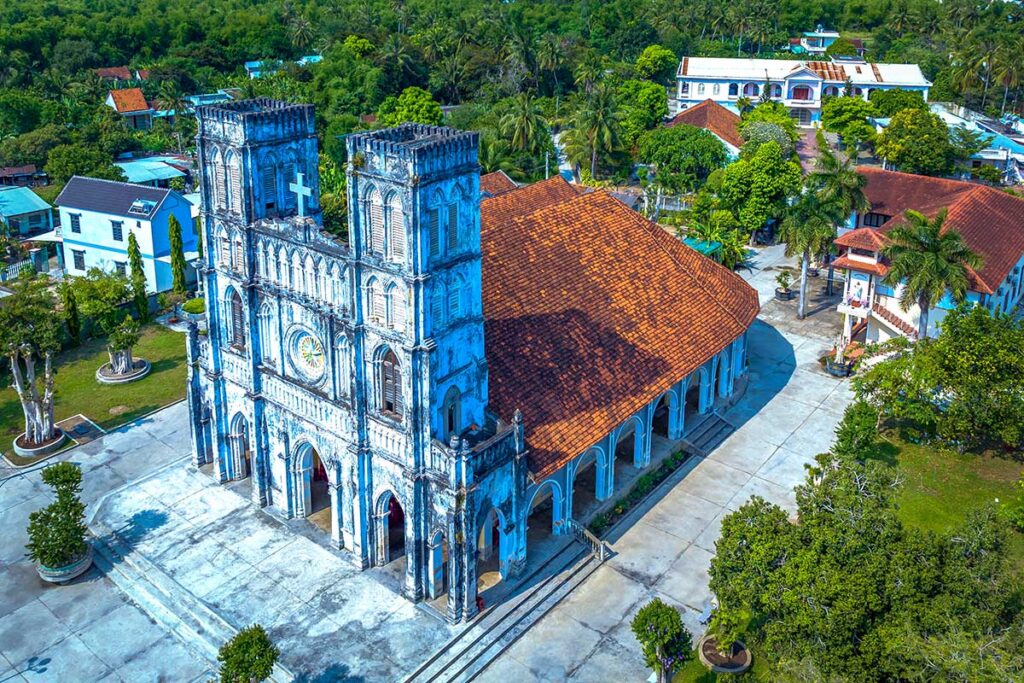 Luftaufnahme der Mang Lang Kirche in Phu Yen, einer gotischen französischen Kirche aus dem neunzehnden Jahrhundert mit blauen Fassaden und roten Dachziegeln, umgeben von Palmen und einem Dorf — eine der ältesten Kirchen Zentralvietnams