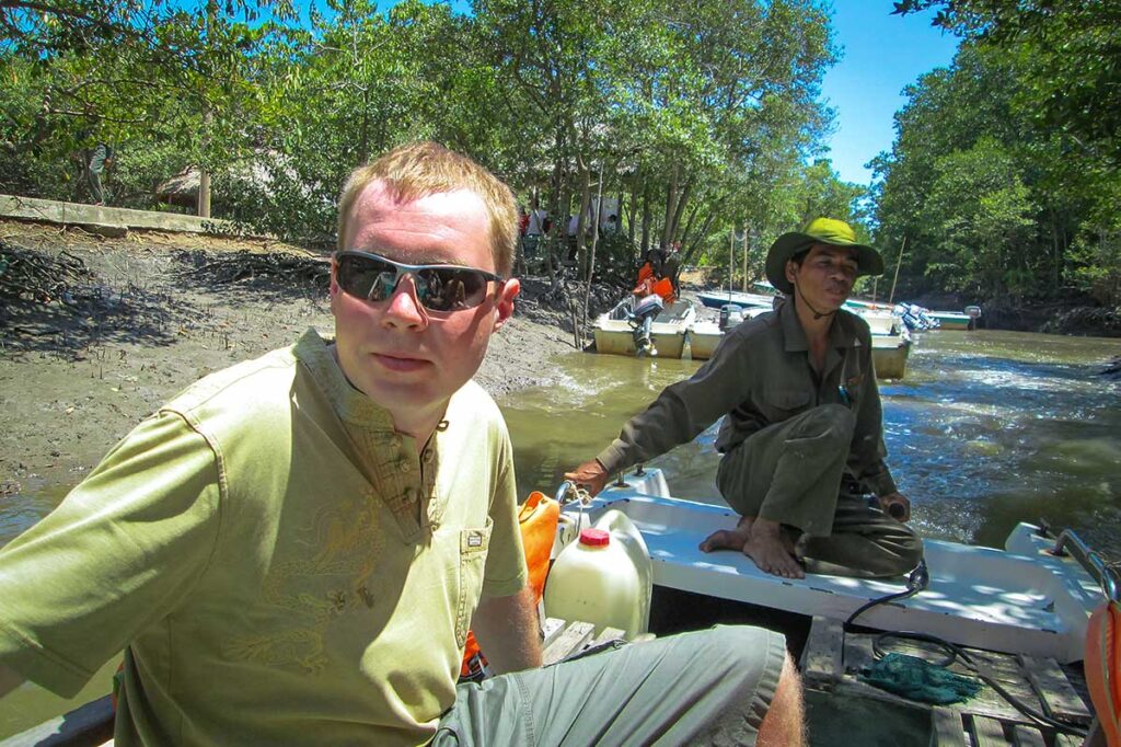 Tourist und lokaler Guide auf einem Motorboot während einer Bootsfahrt durch das Can Gio Reservat, mit Mangrovenbäumen am schlammigen Ufer im Hintergrund — eine Bootsfahrt durch das Reservat ist die gebräuchlichste Art, den Mangrovenwald zu erkunden