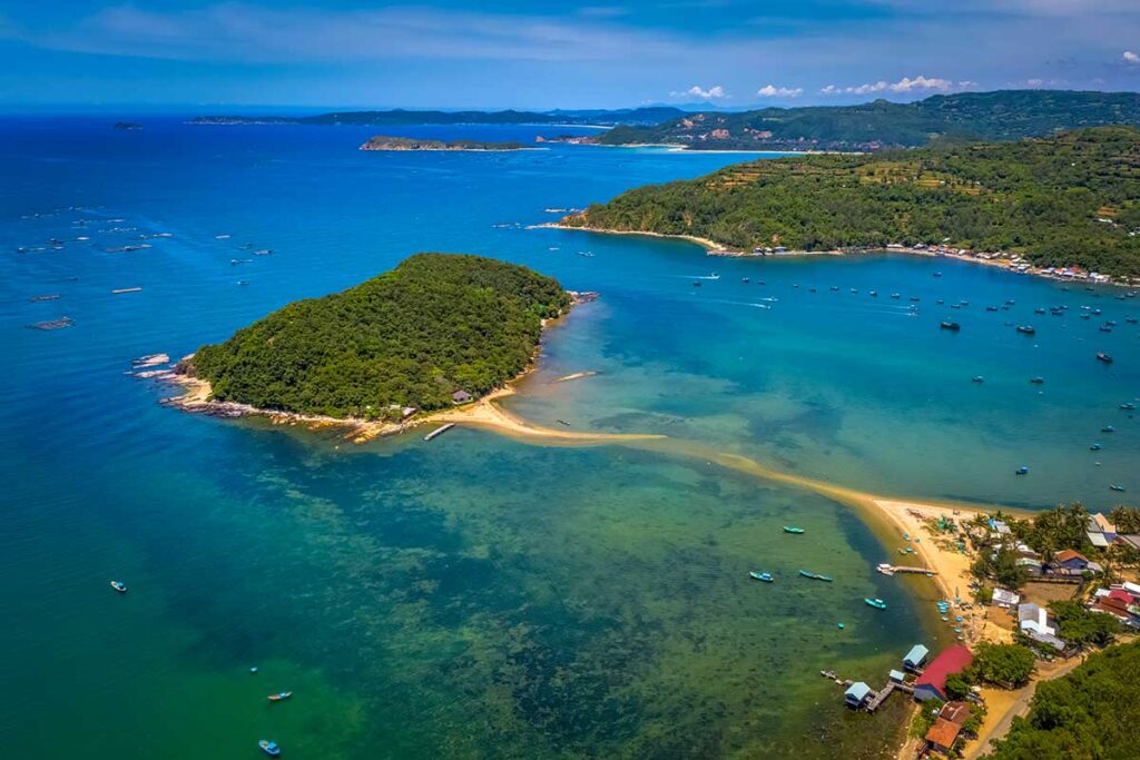 Luftaufnahme der Insel Nhat Tu Son in Phu Yen mit einem bewaldeten Hügel, einer Sandbank, die die Insel mit der Küste verbindet, und Dutzenden Fischerbooten im türkisfarbenen Wasser