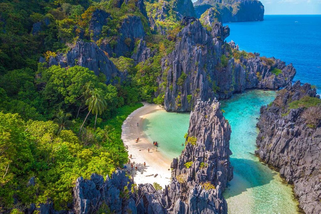 Luftaufnahme eines abgelegenen weißen Sandstrands eingeschlossen von scharfen Kalksteinfelsen und türkisfarbenem Wasser auf Palawan auf den Philippinen, mit einer Handvoll Besuchern am Strand