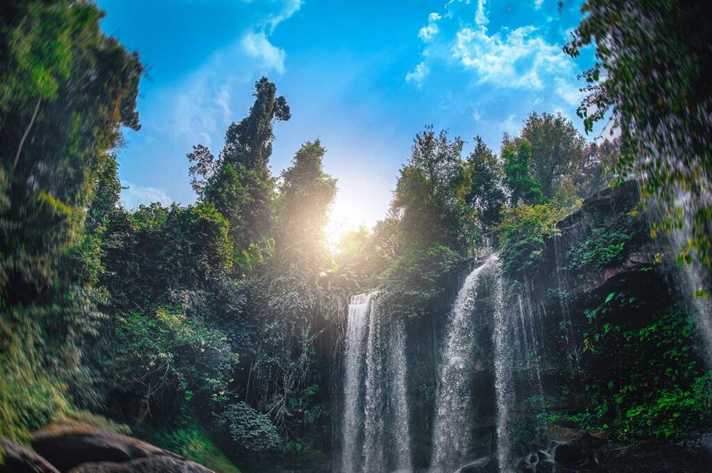 Wasserfall im Dschungelgebiet von Phnom Kulen in Kambodscha, fotografiert aus dem Wasser heraus mit Sonnenlicht das durch die Baumkronen bricht