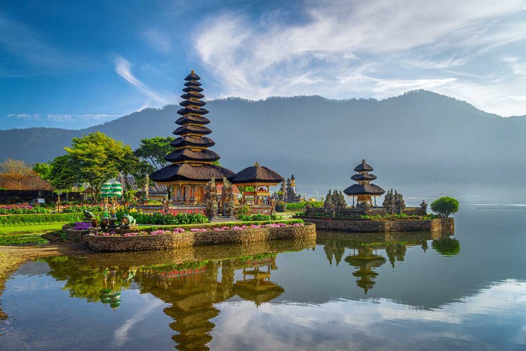 Der Wassertempel Pura Ulun Danu Bratan auf Bali spiegelt sich im ruhigen Seewasser, umgeben von Blumen und Bergen im Nebel