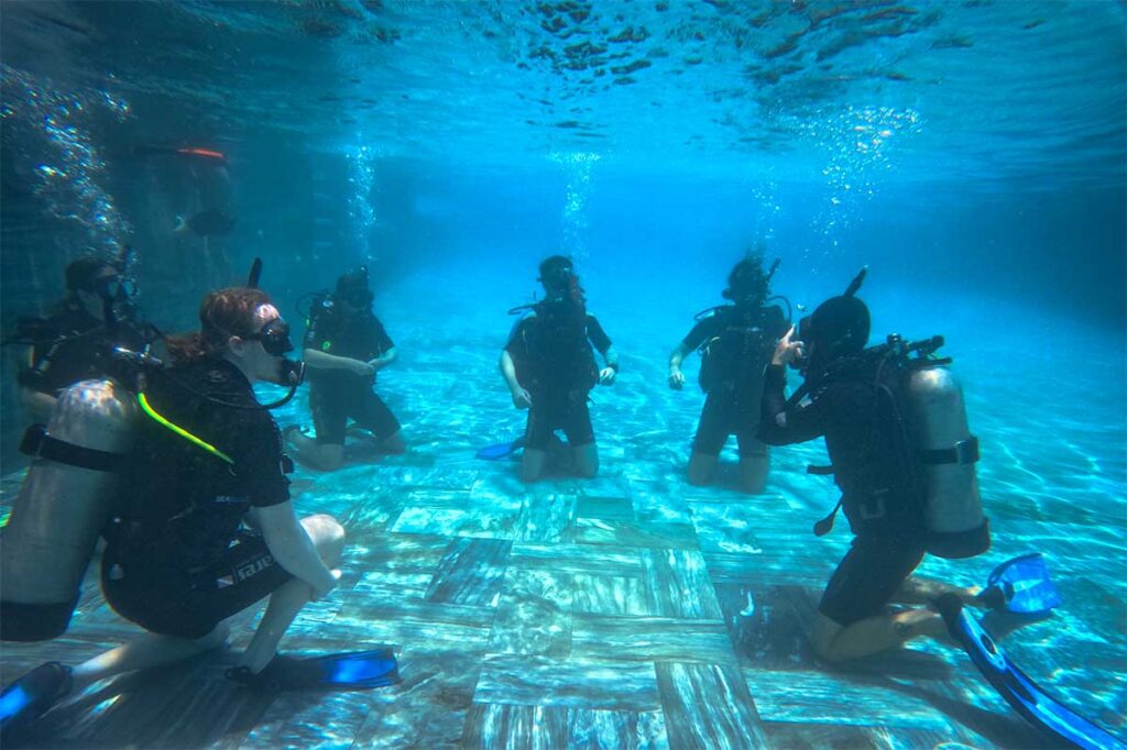 Gruppe von Kursteilnehmern übt Tauchtechniken auf dem Boden eines Schwimmbades während eines PADI-Tauchkurses in Hoi An — das Schwimmbadtraining ist der erste Schritt für alle, die bei Hoi An tauchen lernen möchten