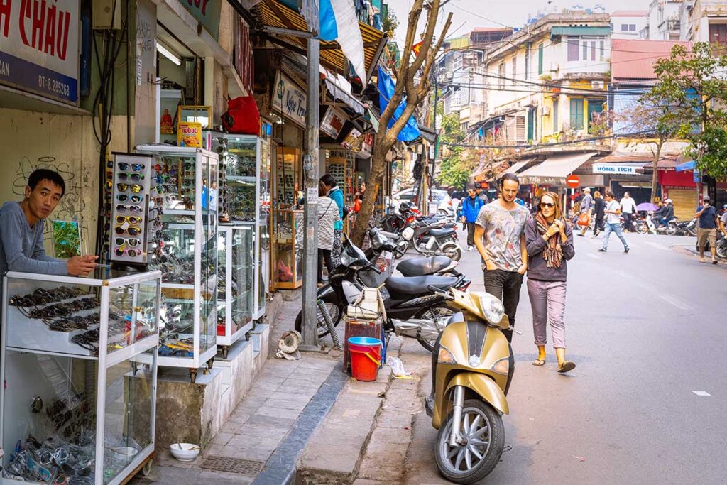 Westliche Touristen gehen an einem Straßenverkäufer mit Sonnenbrillen in der Altstadt von Hanoi, Vietnam, vorbei