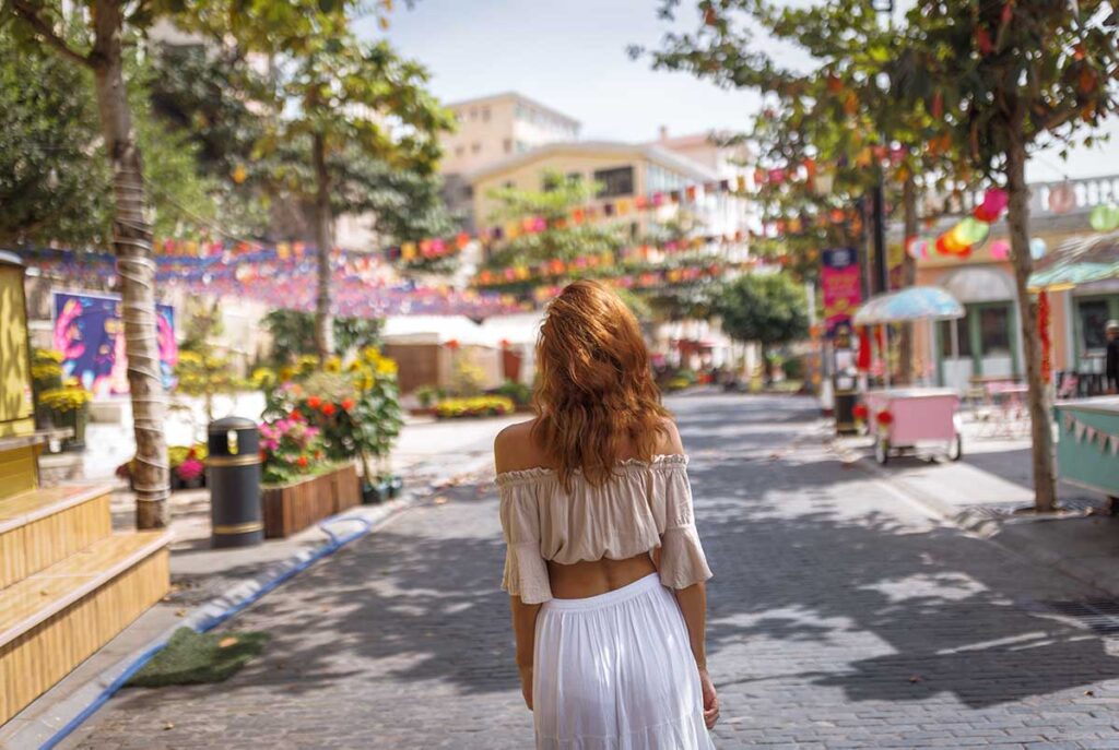 Frau in weißem schulterfreiem Outfit spaziert durch eine sonnige Straße in Vietnam mit bunten Dekorationen und tropischen Bäumen