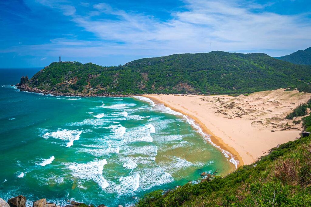 Verlassener Sandstrand von Bai Mon Beach bei Mui Dien in Phu Yen mit kräftigen Wellen, Sanddünen und einem Leuchtturm auf dem bewaldeten Kap — der östlichste Punkt des vietnamesischen Festlands