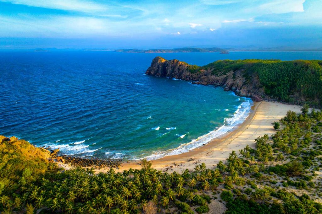 Verlassener halbkreisförmiger Strand in Phu Yen mit goldgelbem Sand, blauen Wellen, Palmen und einem felsigen Kap im Hintergrund — einer der wenig bekannten Strände entlang der Küste von Phu Yen