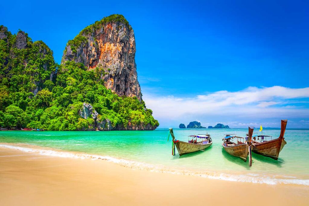 Drei traditionelle Longtailboote im klaren türkisfarbenen Wasser von Railay Beach in Thailand, mit einem großen bewachsenen Kalksteinfelsen im Hintergrund