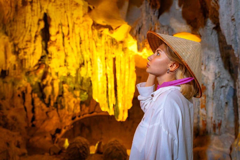 Westlicher Tourist mit vietnamesischem Kegelstrohhut bewundert die Stalaktiten in einer beleuchteten Höhle in Vietnam, verantwortungsvolles Reisen bedeutet auch das Respektieren natürlicher und kultureller Sehenswürdigkeiten
