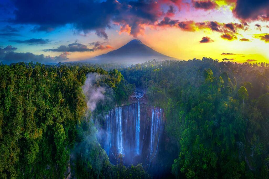Luftaufnahme des Tumpak-Sewu-Wasserfalls auf Java in Indonesien, der in eine bewaldete Schlucht stürzt, mit dem konischen Gipfel des Semeru-Vulkans im Hintergrund bei Sonnenuntergang