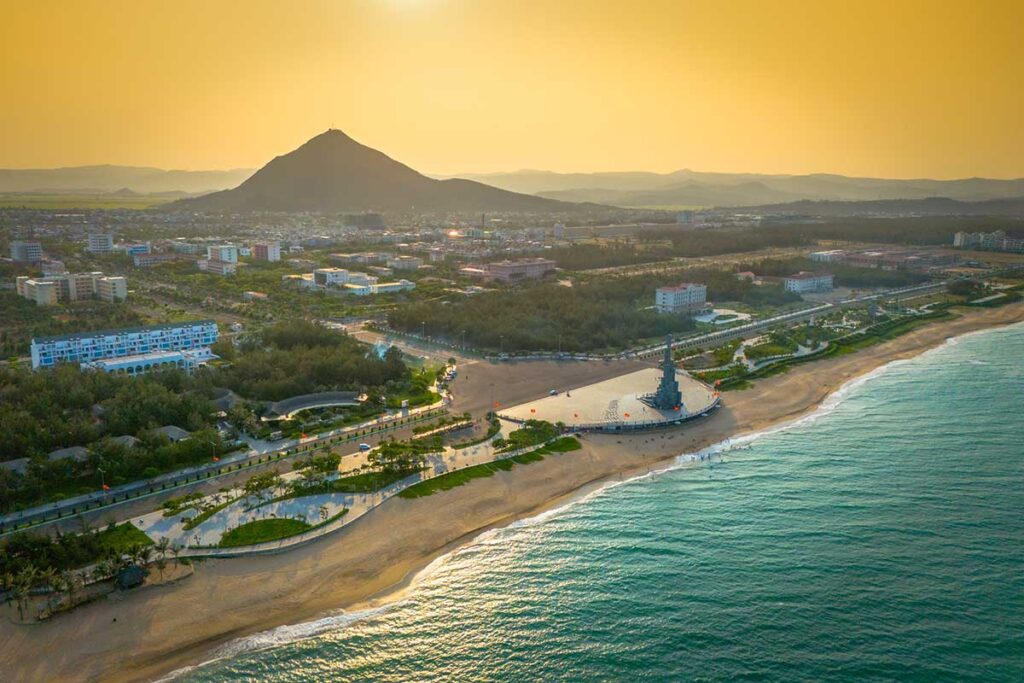 Luftaufnahme der Küstenlinie von Tuy Hoa bei Sonnenuntergang mit dem Nghinh Phong Turm am Strand, einer langen Strandpromenade, Stadtgebäuden und einem konischen Berg im Hintergrund