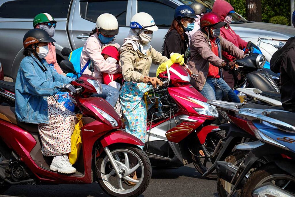 Vietnamesische Motorradfahrer an einer belebten Kreuzung mit vollständigem Sonnenschutz, darunter Gesichtsmasken, Handschuhe und lange Ärmel