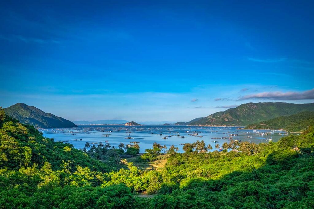 Panoramablick über die Vung Ro Bay in Phu Yen mit klarem blauem Wasser, Fischzuchtanlagen und Fischerbooten verteilt über die Bucht, umgeben von dicht bewaldeten Hügeln — eine abgelegene Bucht nahe der Grenze zu Khanh Hoa