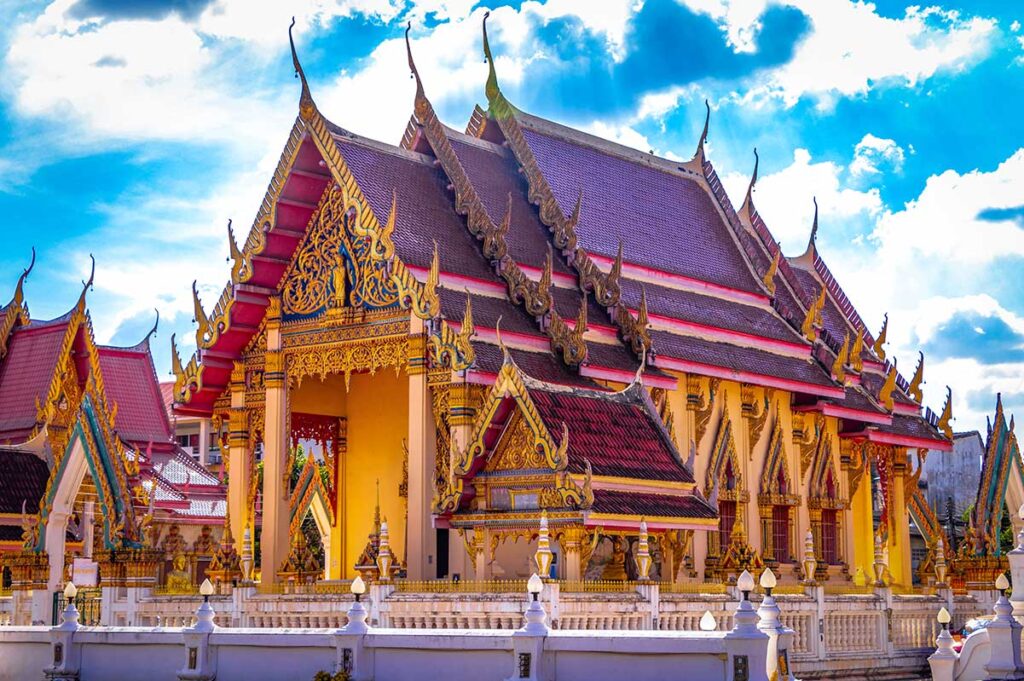 Thailändischer buddhistischer Tempel mit goldenen Ornamenten, rotem Dach und weißen Mauern unter blauem Himmel mit Wolken