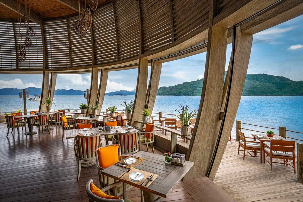 Offenes Restaurant des An Lam Retreats Ninh Van Bay mit Holzstühlen mit orangefarbenen Kissen, Panoramafenstern mit Blick über die blaue Bucht und Inseln in der Ferne