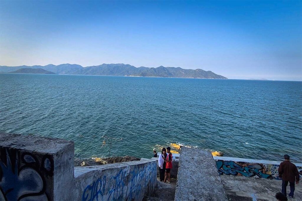 Aussichtspunkt an der Küste der Ninh Van Bay mit Besuchern auf Betontreppen mit Graffiti, Blick über das dunkelblauwe Meer und den bewaldeten Bergrücken von Ninh Van im Hintergrund
