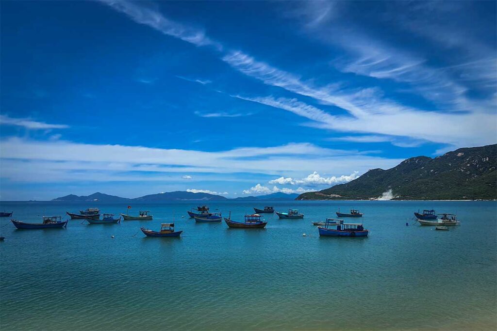 Dutzende blaue Fischerboote und runde Korbboote liegen vor Anker in der ruhigen Bucht von Ninh Van, mit grünen Hügeln und einer felsigen Insel im Hintergrund — das Herz dieses abgelegenen Fischerdorfs