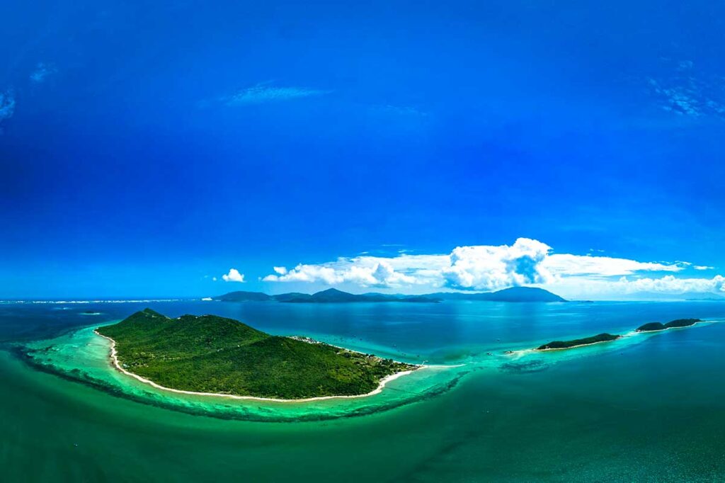 Panoramische Luftaufnahme der drei Inseln von Diep Son in der Van Phong Bay, umgeben von hellblauem und türkisfarbenem Wasser mit unter der Oberfläche sichtbaren Korallenriffen und Bergen am Horizont
