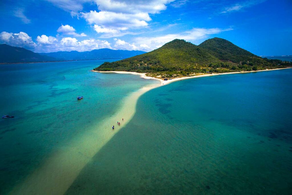 Luftaufnahme der schmalen Sandbank von Diep Son Insel in der Van Phong Bay, mit Besuchern, die über den flachen Sand zwischen zwei Abschnitten türkisfarbenen Wassers laufen und einem bewaldeten Hügel im Hintergrund