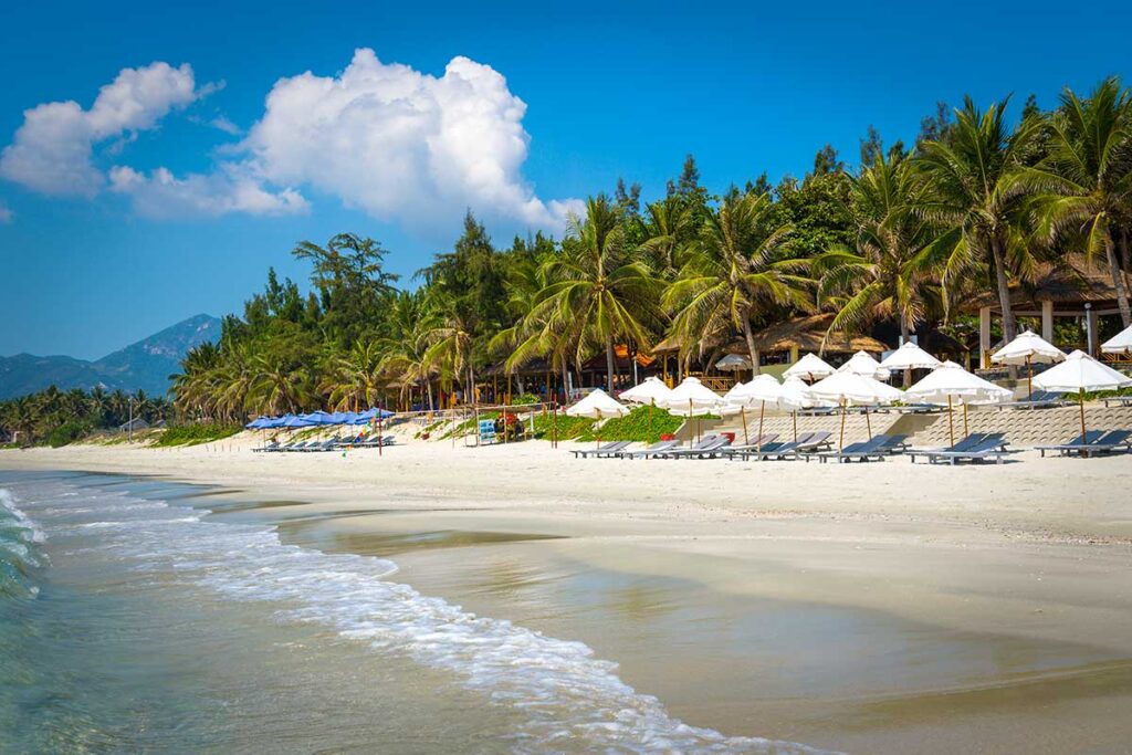 Breiter weißer Strand bei Doc Let Beach mit Reihen von Liegestühlen und weißen Sonnenschirmen vor einem Resort, Palmen und Kiefern im Hintergrund und leichten Wellen an der Wasserlinie