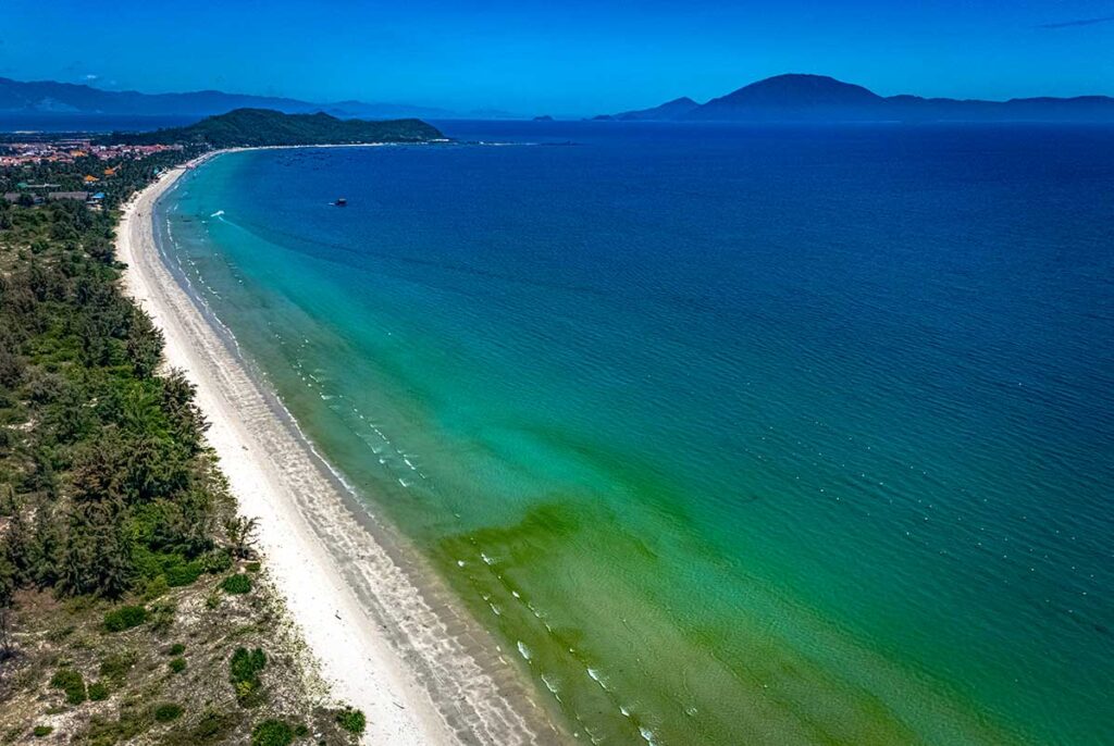 Luftaufnahme des langen weißen Strandes von Doc Let Beach mit klarem grünem und blauem Wasser, einer bewaldeten Küstenlinie und Bergen am Horizont — einer der längsten unberührten Strände bei Nha Trang