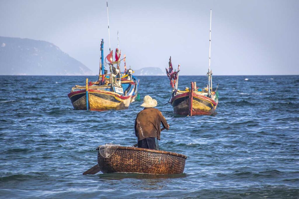 Vietnamesischer Fischer paddelt in einem traditionellen runden Korbboot zu größeren Holzfischerbooten auf See bei Doc Let Beach — ein authentisches Bild des Alltagslebens entlang dieser Küste