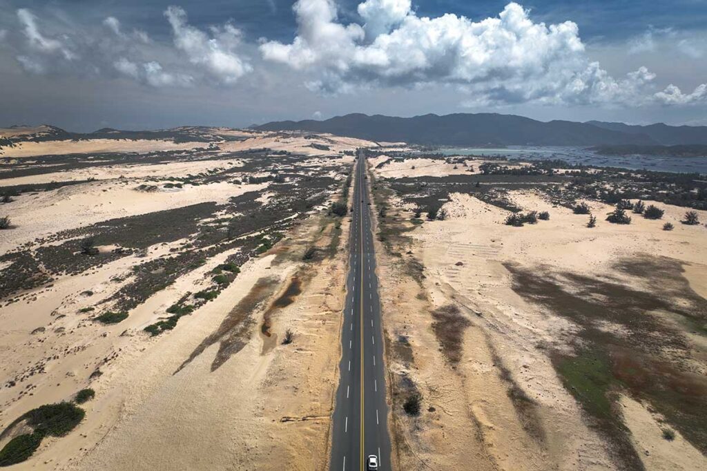 Gerade Straße durch die kahlen Sanddünen der Van Phong Bay mit der Bucht und Bergen am Horizont — eine unerwartet wüstenartige Landschaft an der Küste Zentralvietnams