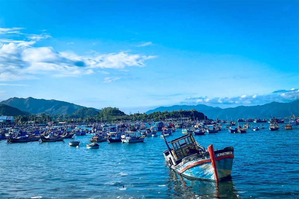 Belebter Fischerhafen der Ninh Van Bay mit Hunderten blauer Fischerboote auf dem Wasser, einem verwitterten Boot im Vordergrund und bewaldeten Hügeln im Hintergrund