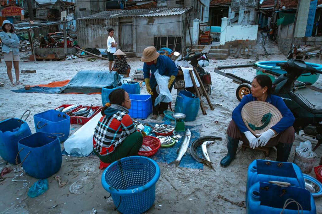 Fischerinnen sortieren und verkaufen frischen Fisch am Strand von Ninh Thuy bei Doc Let, umgeben von blauen Körben und Kisten — ein alltägliches Bild in diesem aktiven Fischerdorf