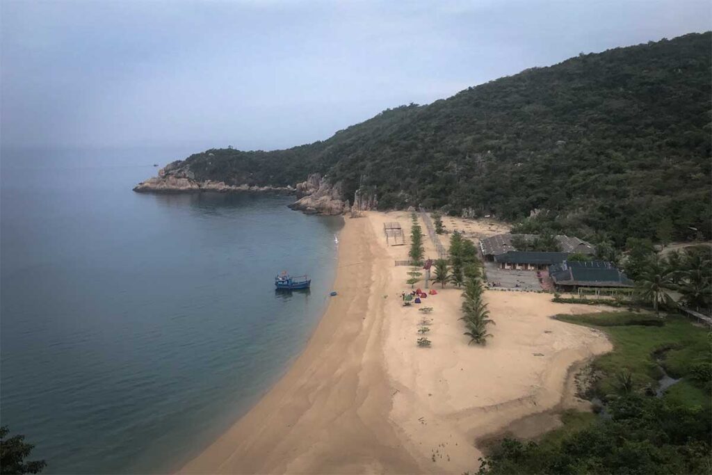 Verlassener goldgelber Strand auf Hon Lon in der Van Phong Bay mit einem Fischerboot im ruhigen Wasser und bewaldeten Hügeln im Hintergrund — eine der wenig erschlossenen Inseln der Bucht