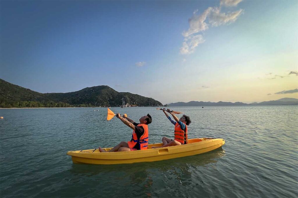 Zwei Kajakfahrer in orangefarbenen Schwimmwesten heben ihre Paddel in einem gelben Kajak auf dem ruhigen Wasser der Ninh Van Bay bei Sonnenuntergang, mit bewaldeten Hügeln und Inseln im Hintergrund