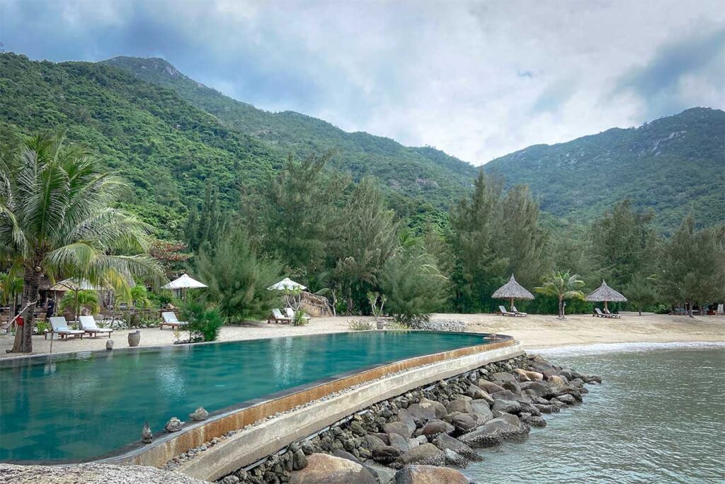 Pool des Lalya Resorts am Rand des Strandes in der Ninh Van Bay, umgeben von Kiefern und grünen Berghängen — eines der exklusiven Resorts, die nur per Boot erreichbar sind