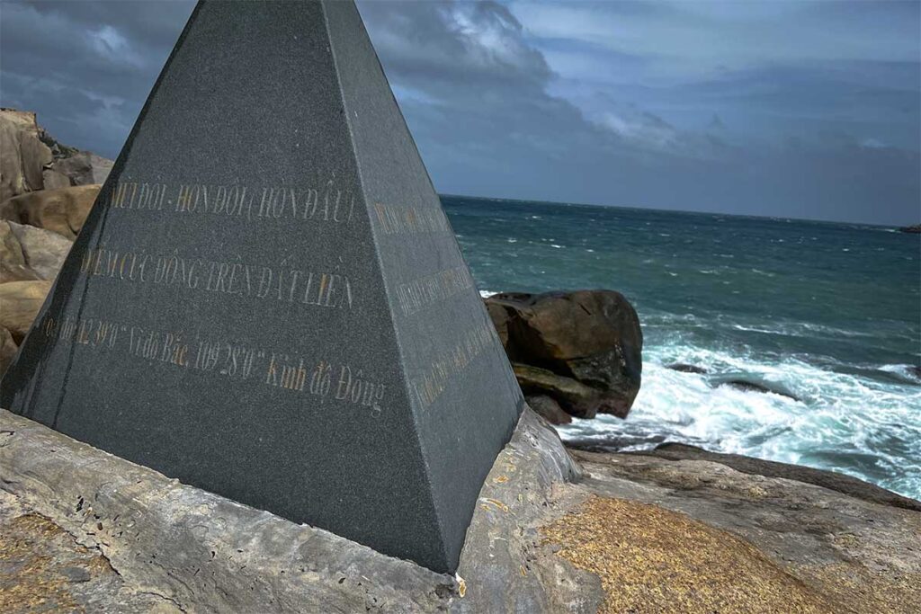 Granit-Gedenkstein auf Mui Doi mit vietnamesischer Inschrift, die diesen Ort als östlichsten Punkt des vietnamesischen Festlands ausweist, mit rauer See im Hintergrund