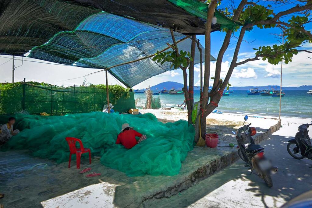 Frau repariert ein großes grünes Fischnetz im Fischerdorf Ninh Thuy bei Doc Let Beach, mit Fischerbooten auf dem Wasser im Hintergrund — ein alltägliches Bild in dieser aktiven Fischergemeinde