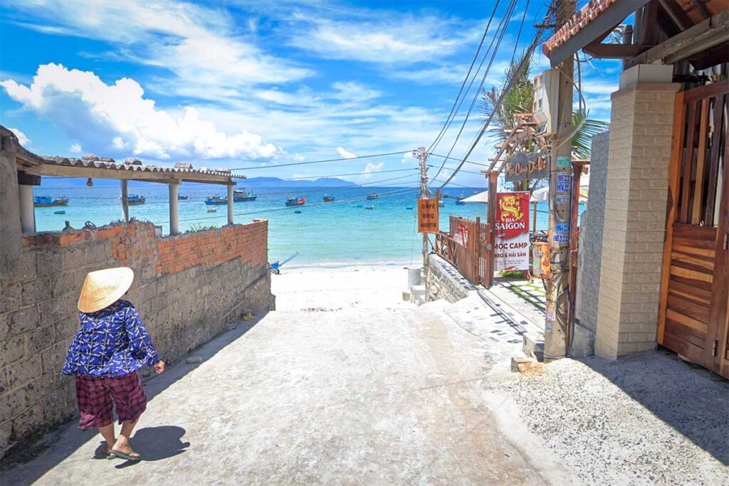 Schmalle Betongasse im Fischerdorf Ninh Thuy mit Blick auf das türkisfarbene Wasser von Doc Let Beach, Fischerbooten in der Bucht und lokalen Gastronomiaschildern am Wegesrand