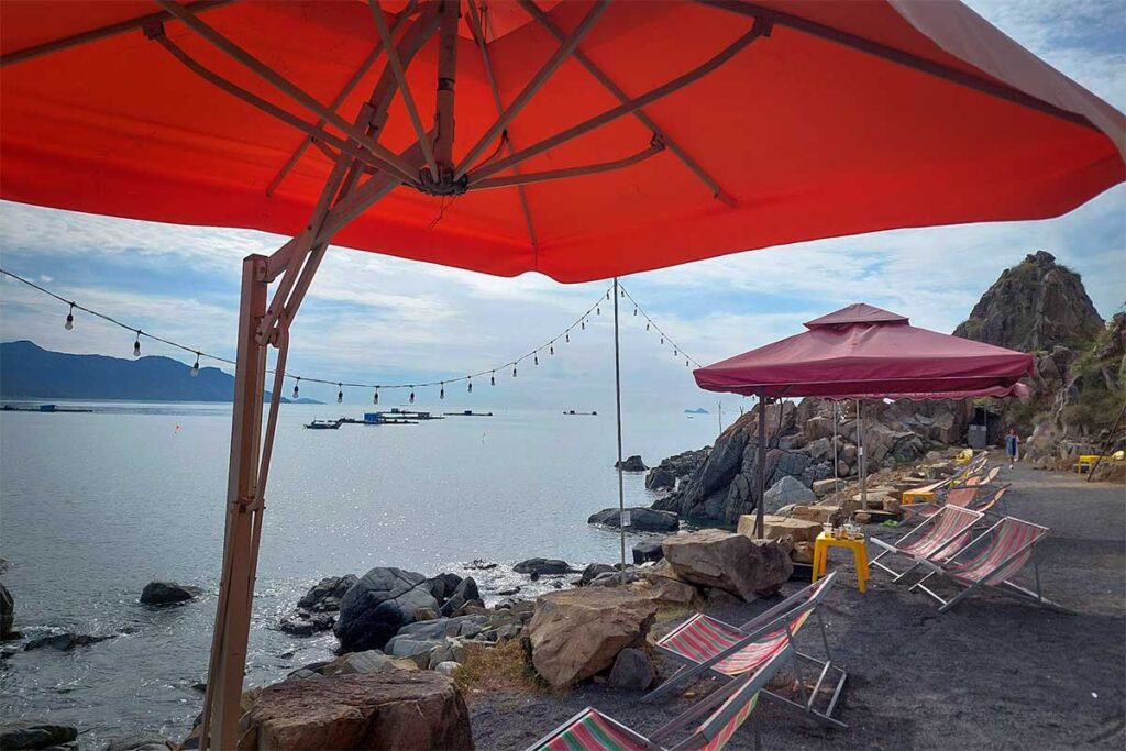 Aussichtscafe an der Felsenküste der Ninh Van Bay mit roten Sonnenschirmen, Liegestühlen und einem Panoramablick auf die Bucht — ein informeller Ort zum Erkunden der Umgebung