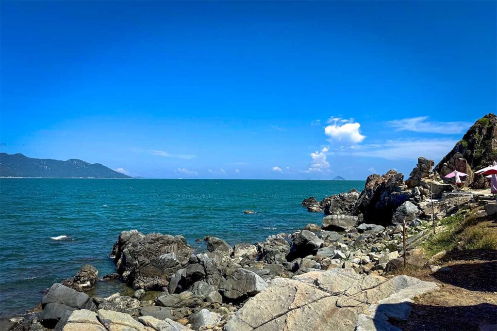 Felsküste der Ninh Van Bay mit großen dunklen Steinbrocken, türkisfarbenem Wasser und einem kleinen Cafe mit Sonnenschirmen im Hintergrund — die raue Küstenlinie macht Ninh Van Bay einzigartig entlang der Küste von Nha Trang